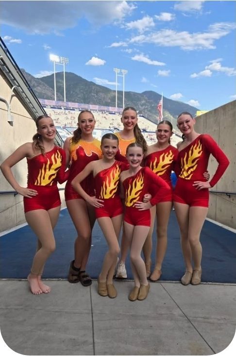 Group of dancers in red and gold costumes posing outdoors, smiling. Mountain backdrop.
