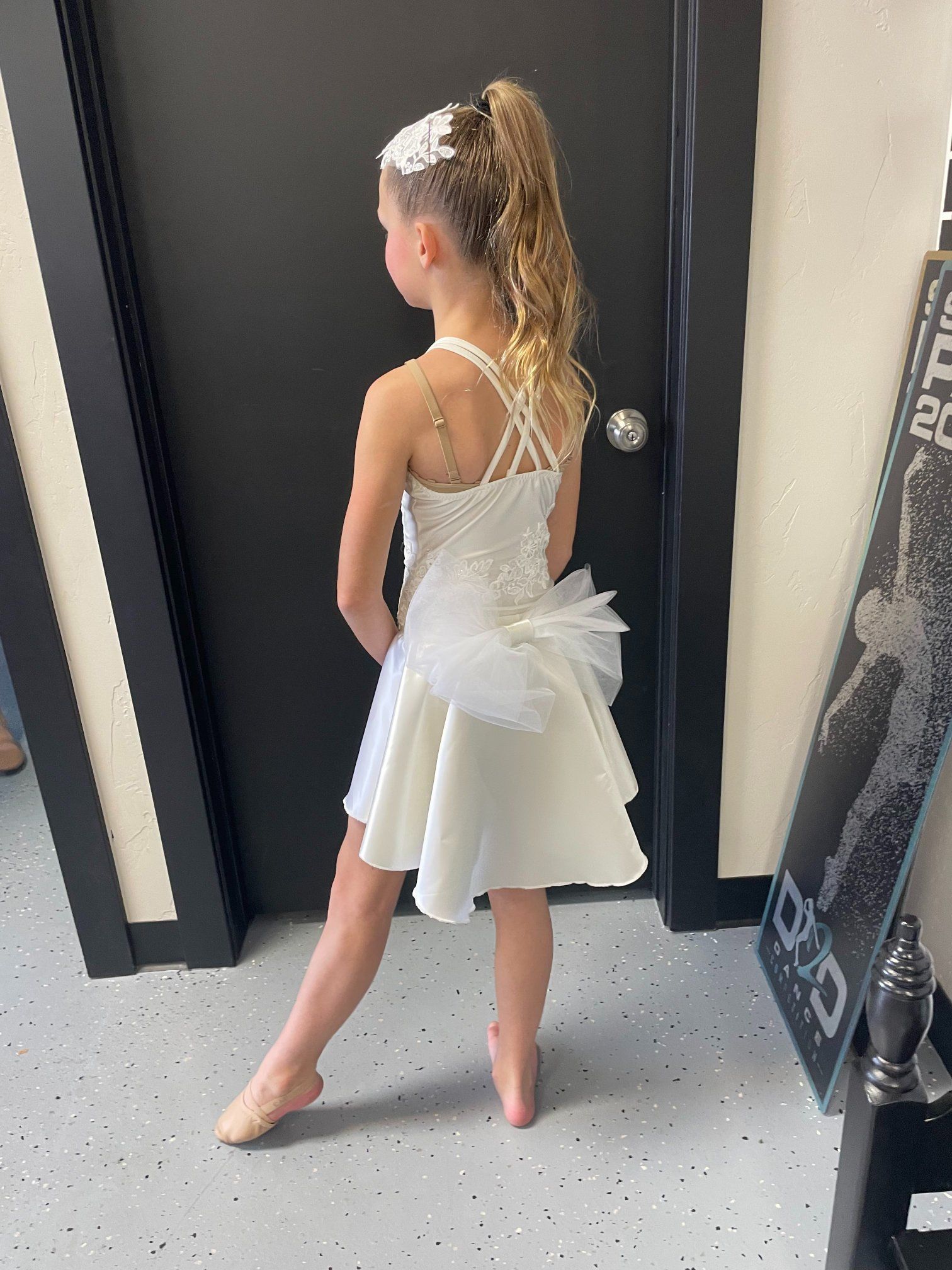 Girl in white dance dress and bun, standing en pointe near a black door, looking away.