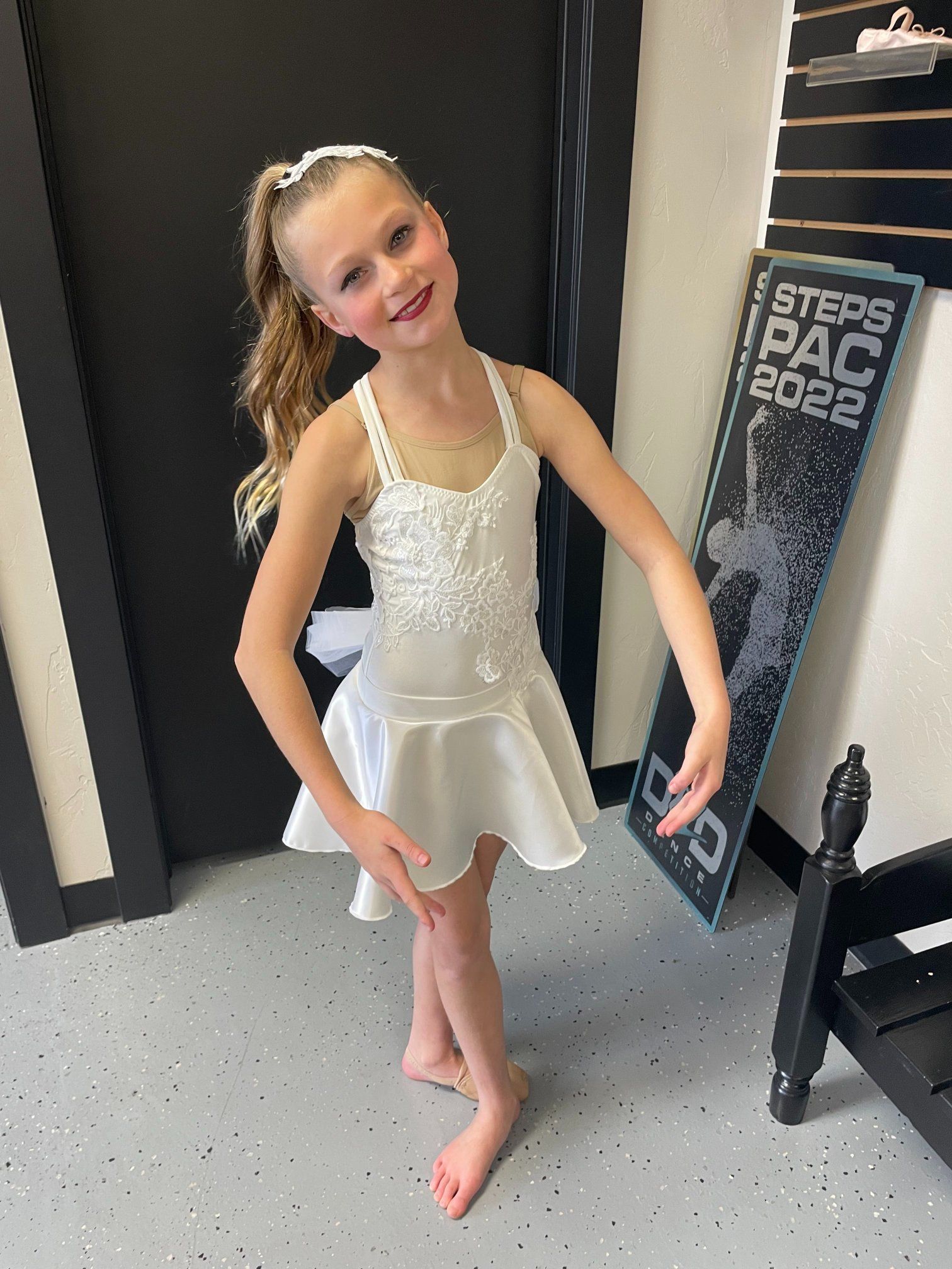 Young girl in a white dance costume, smiling, with hands in ballet pose, in a dance studio.