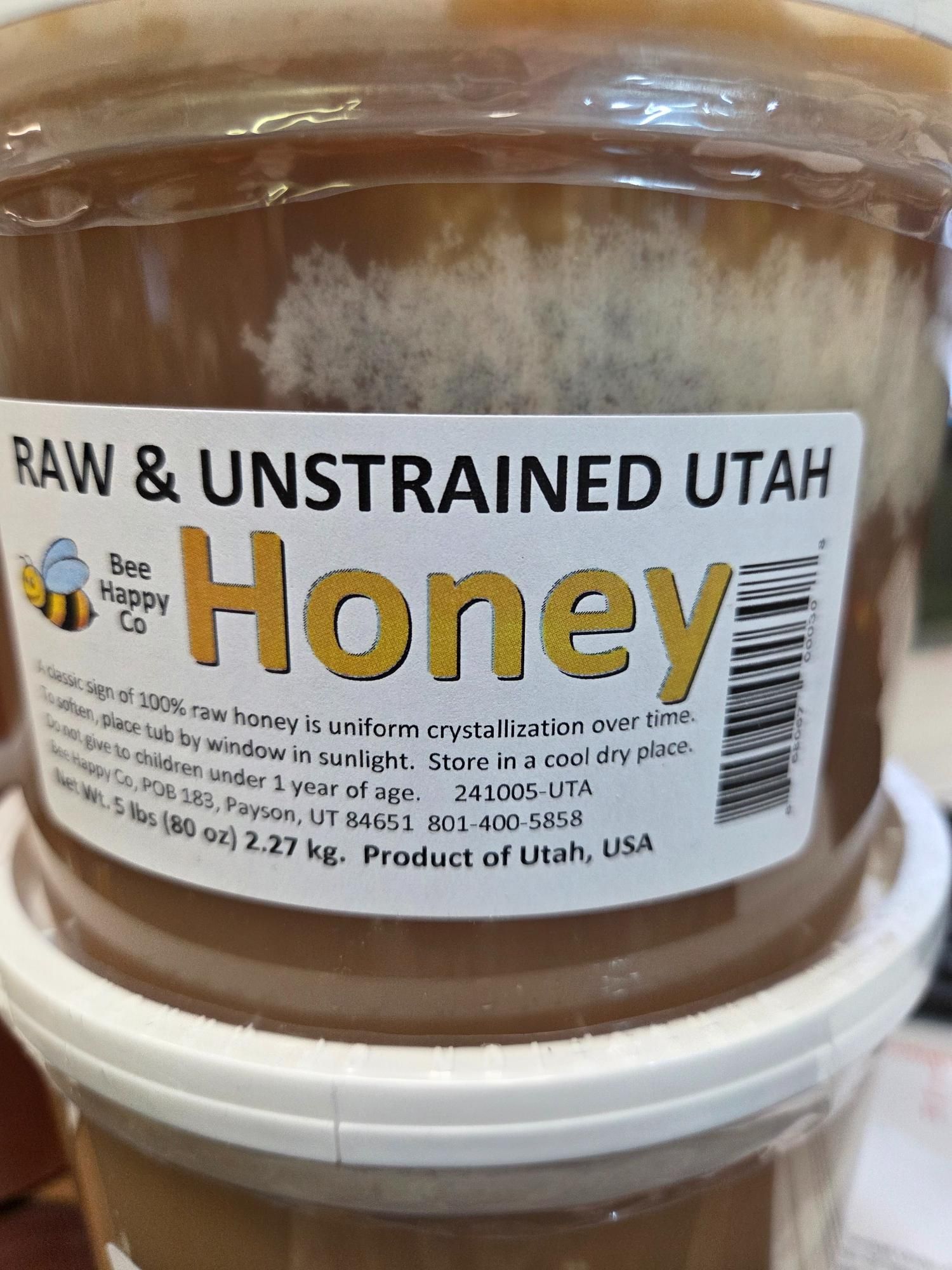 Close-up of a container of raw and unstrained honey from Utah. White label with text, a clear tub with honey.
