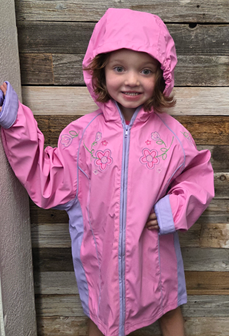 Girl in pink hooded jacket smiles, leaning against a wood wall.