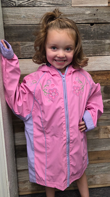 Girl with curly hair in a pink and purple jacket, smiles at the camera. Against a wooden wall.