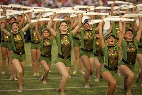 Dancers in green sequined outfits and hats holding white flags, performing on a green field.
