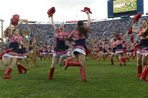 Cheerleaders in red boots and hats, jumping on a football field in front of a crowd at a stadium.