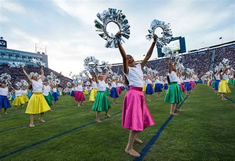 Dancers in colorful skirts and white shirts perform on a green field, holding silver props, stadium in the background.