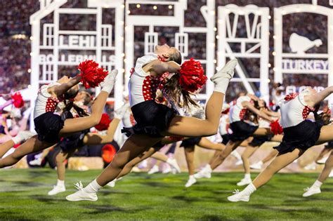Cheerleaders in black skirts, red pom-poms, and white sneakers leap on green grass in front of a large stage.