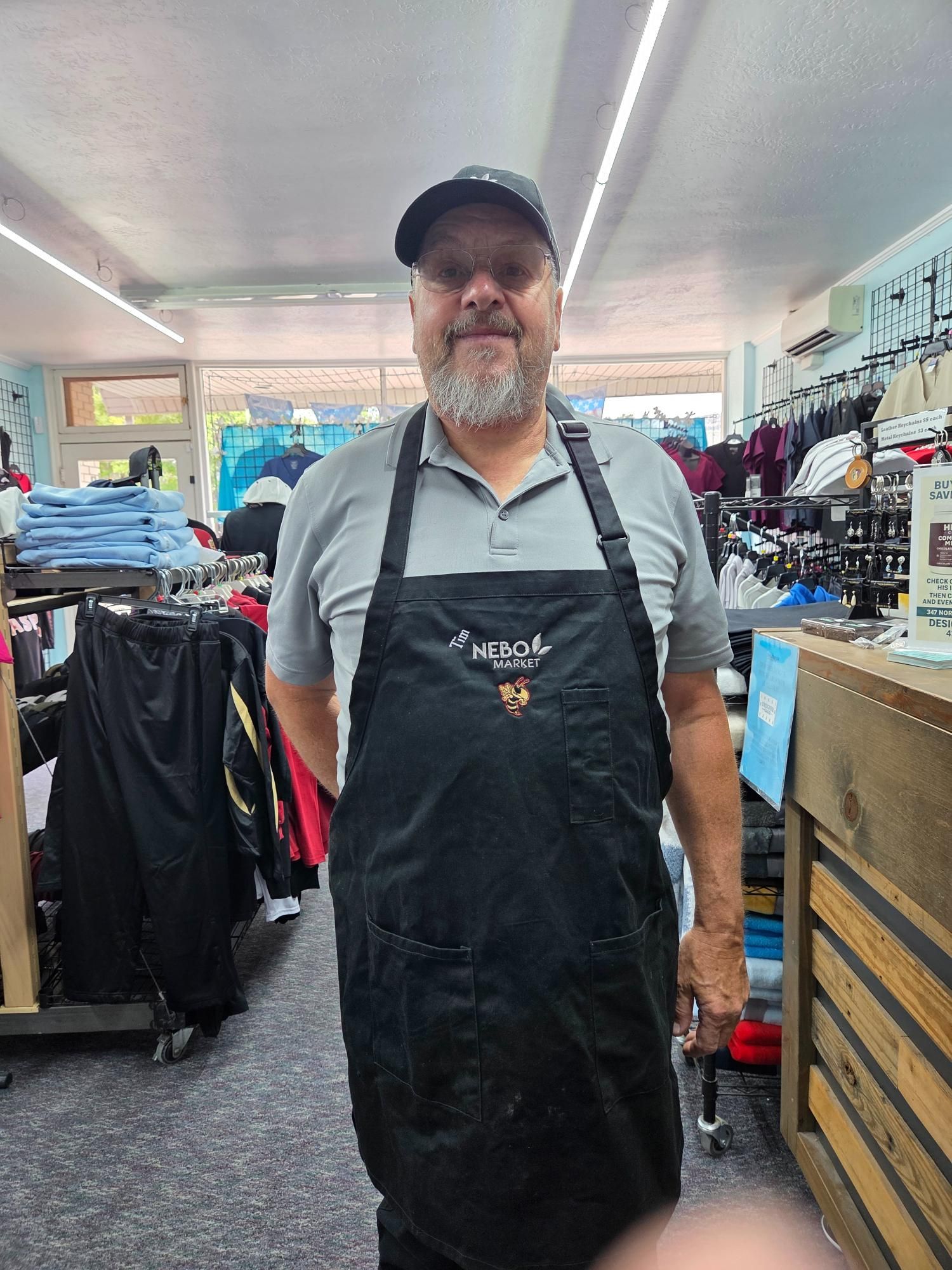 Man in apron and cap standing in a store, smiling. Clothes and shelves are visible in the background.