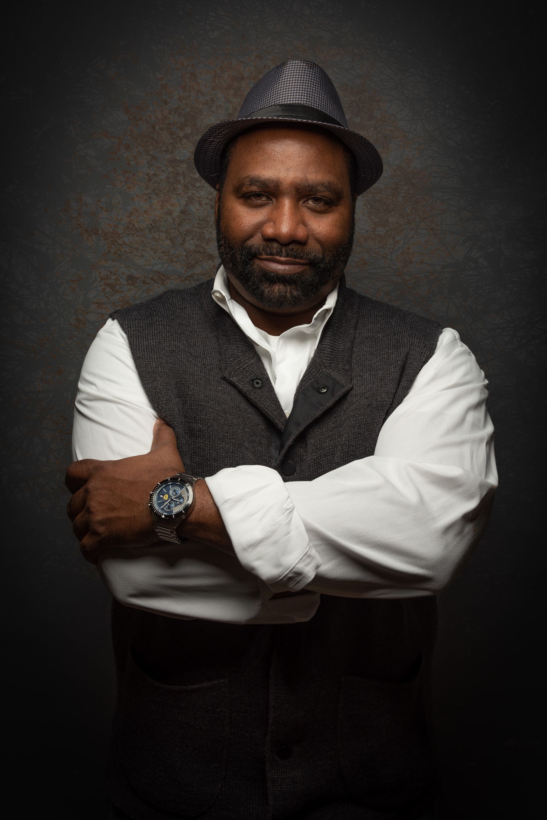 Man with dark skin, wearing a hat, vest, and watch, arms crossed. Dark background.