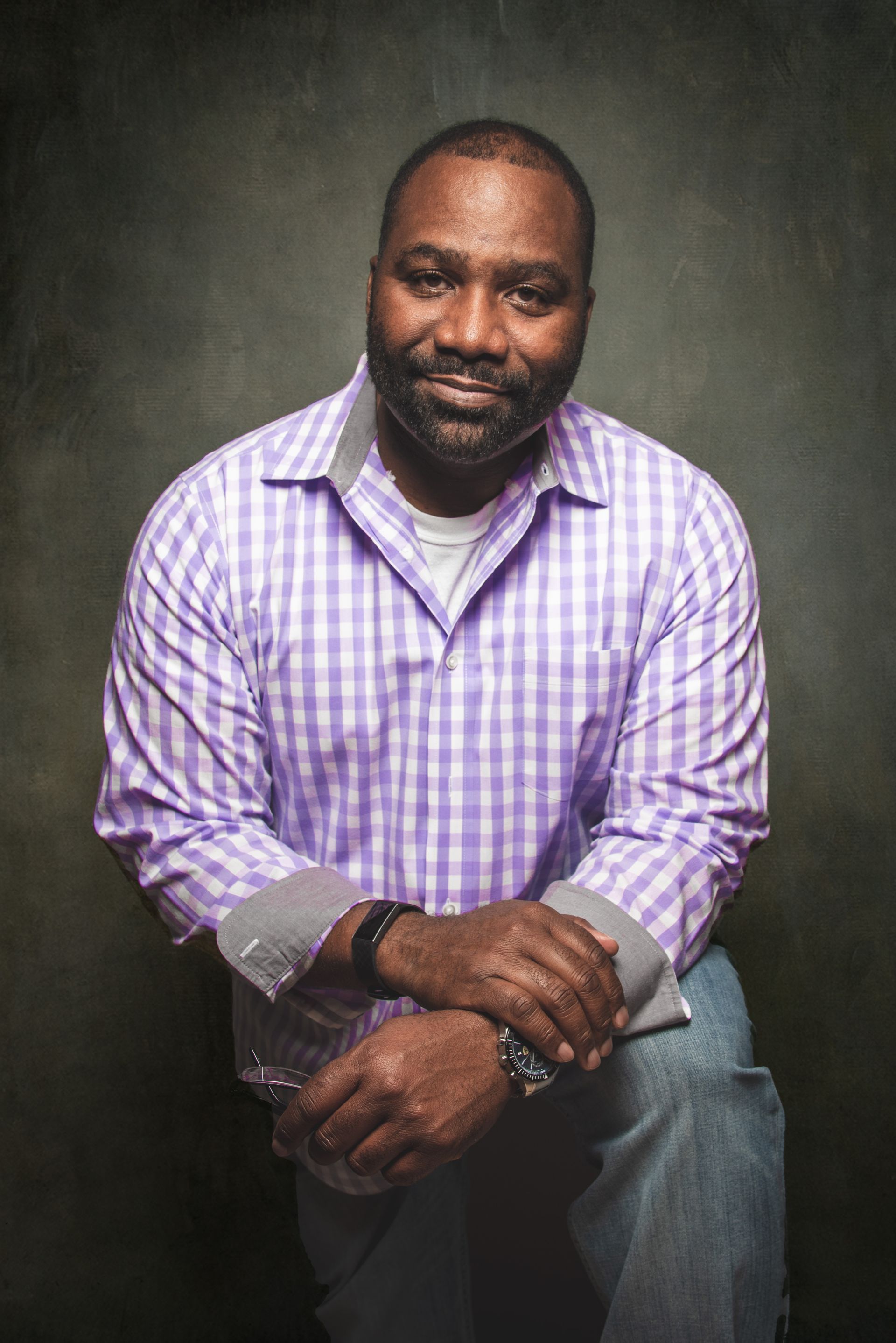 Man with a beard in a purple plaid shirt, jeans, and a gray undershirt, seated.