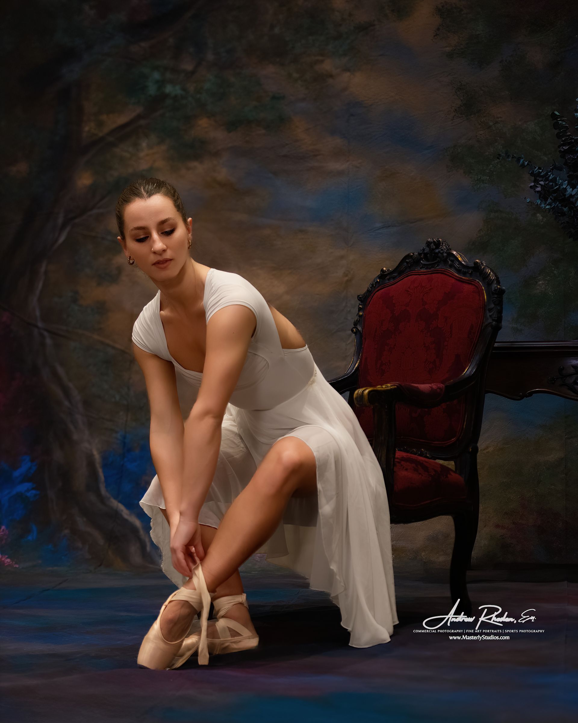 Ballet dancer in white dress, tying pointe shoe, seated near ornate red chair, with painted forest backdrop.