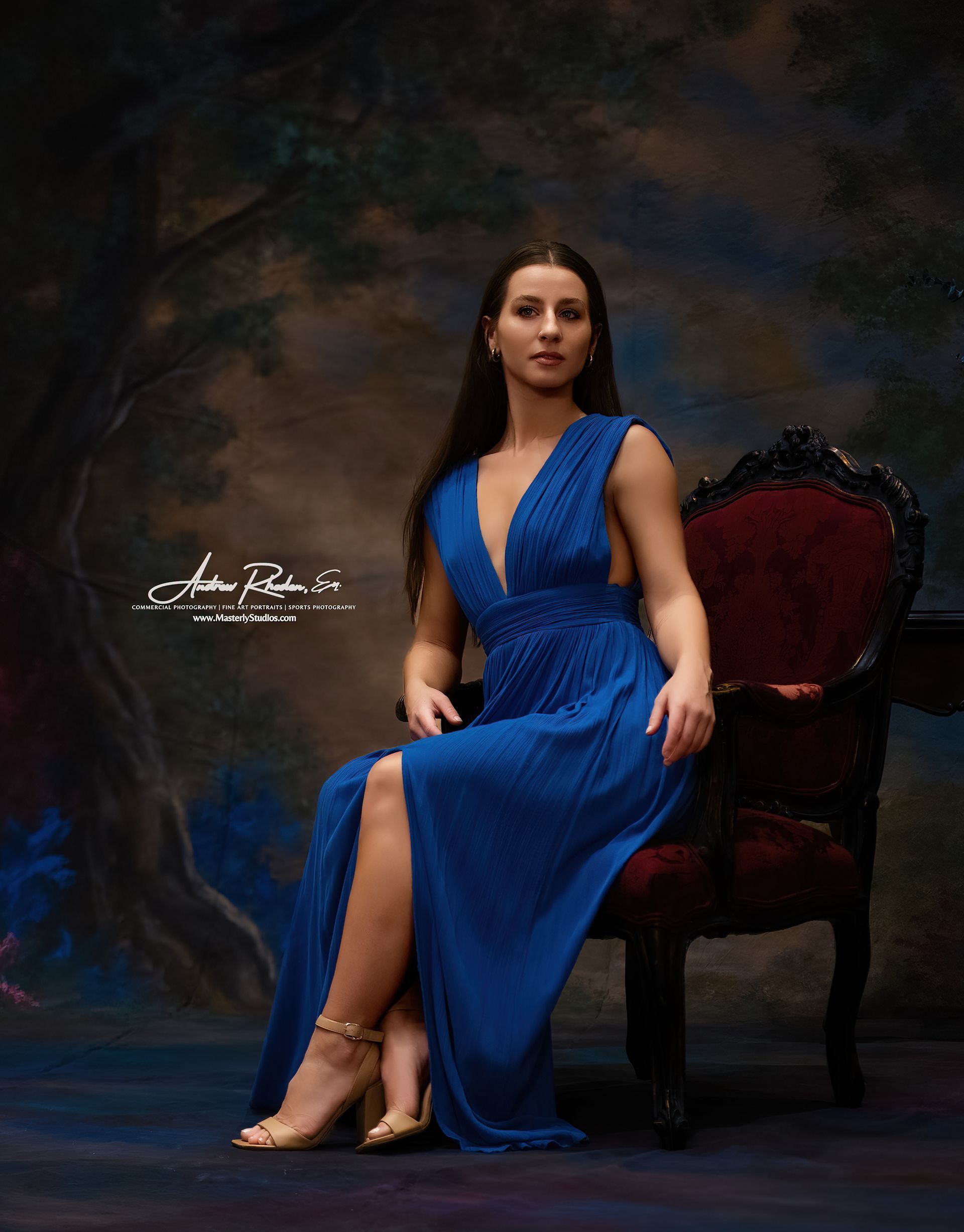 Woman in a blue dress seated in a vintage chair against a dark painted backdrop.
