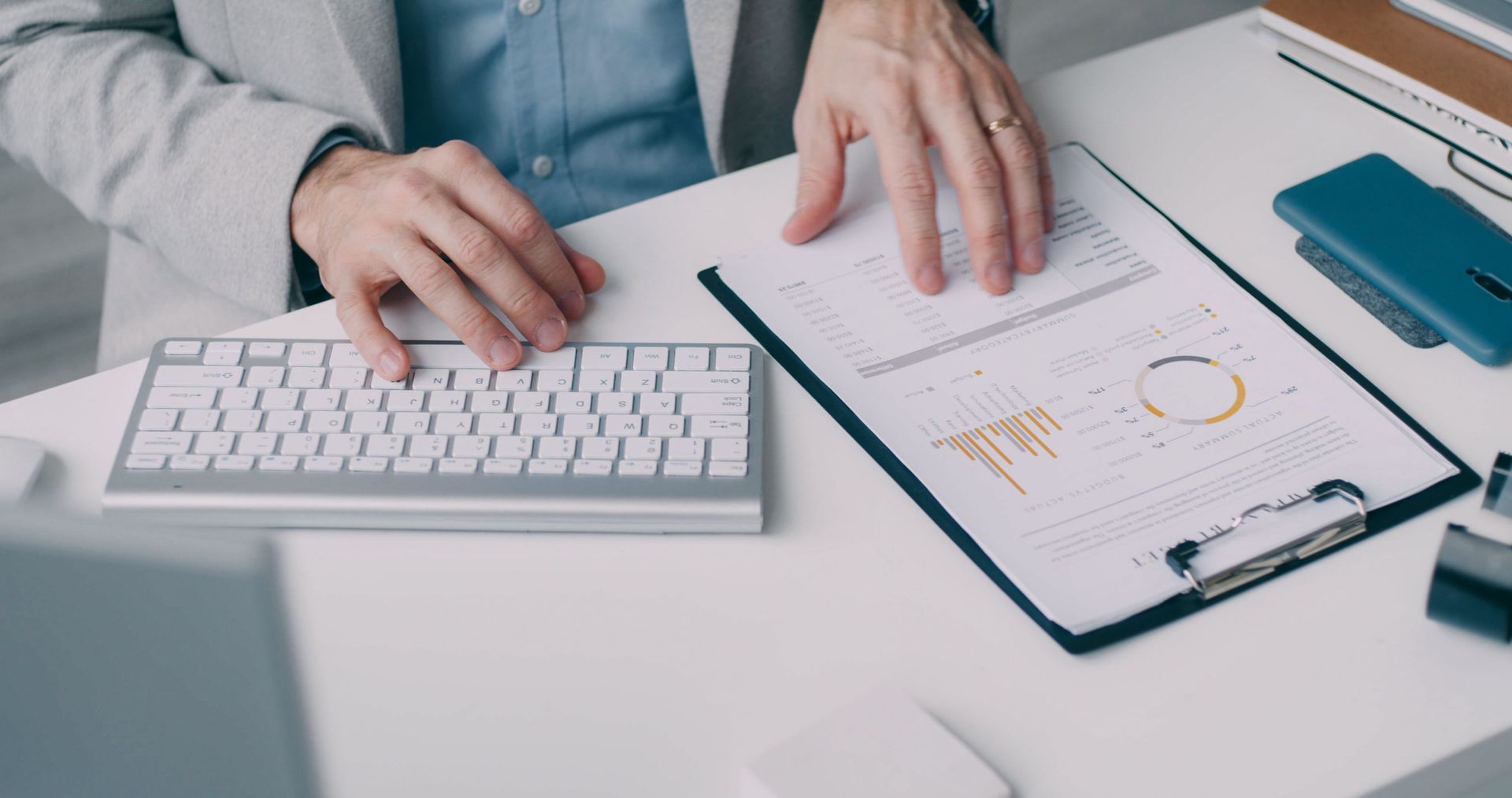 Hands typing on a white keyboard beside a clipboard with charts and a teal phone on a desk