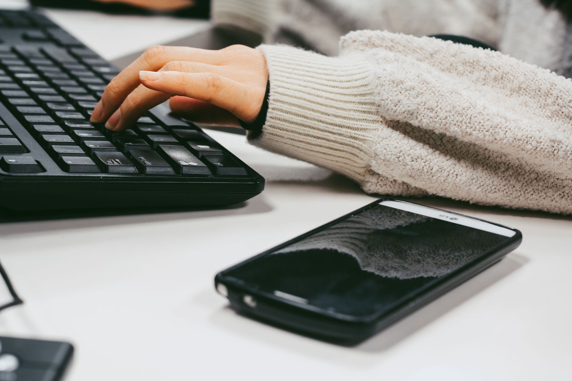 Hand typing on a black keyboard beside a smartphone on a white desk
