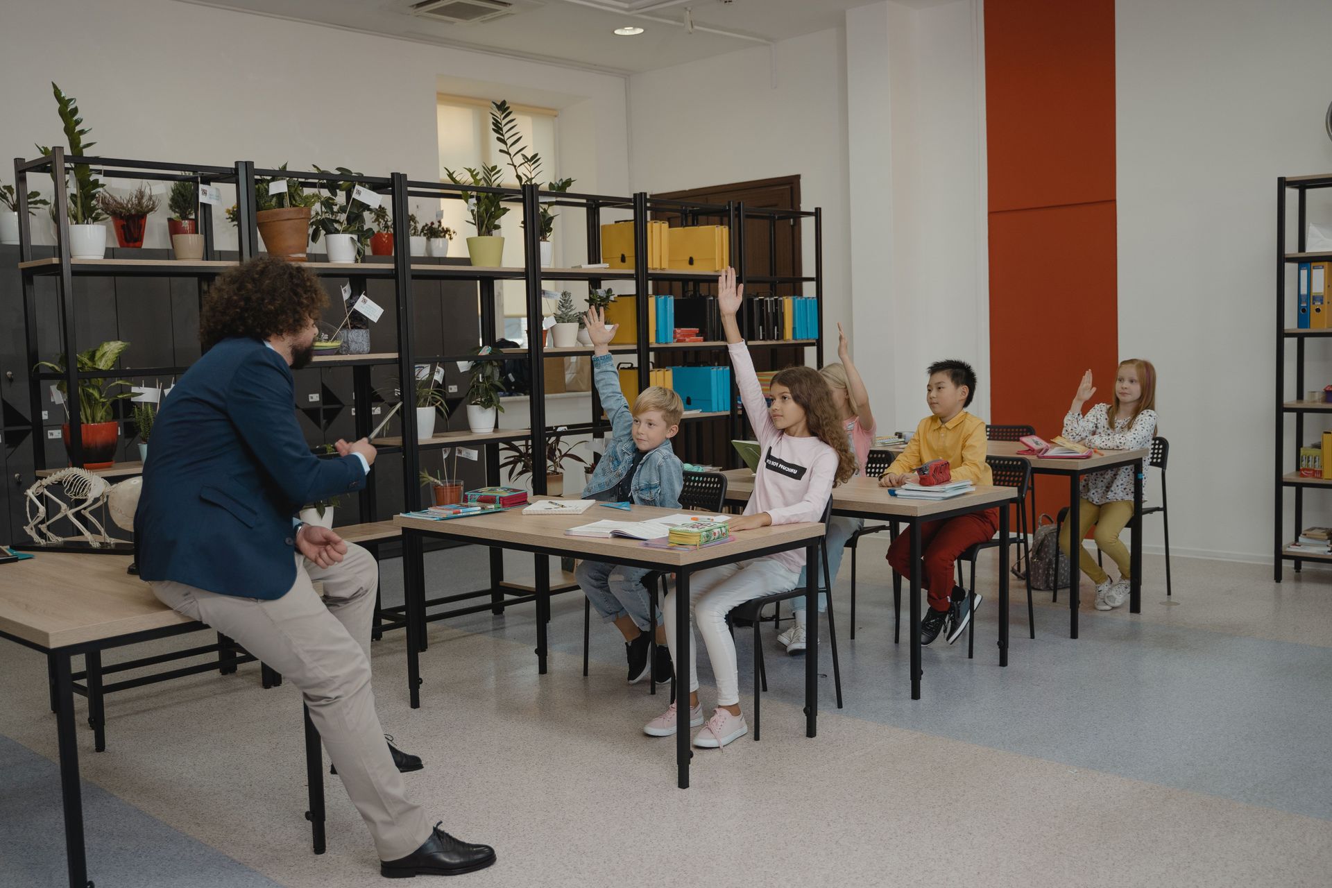Teacher leading seated class in a bright room, with students raising hands at tables