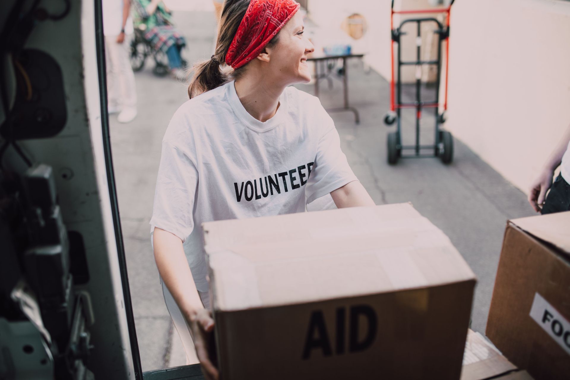 Volunteer in a white shirt and red bandana carrying a box labeled “AID” in a relief setting.