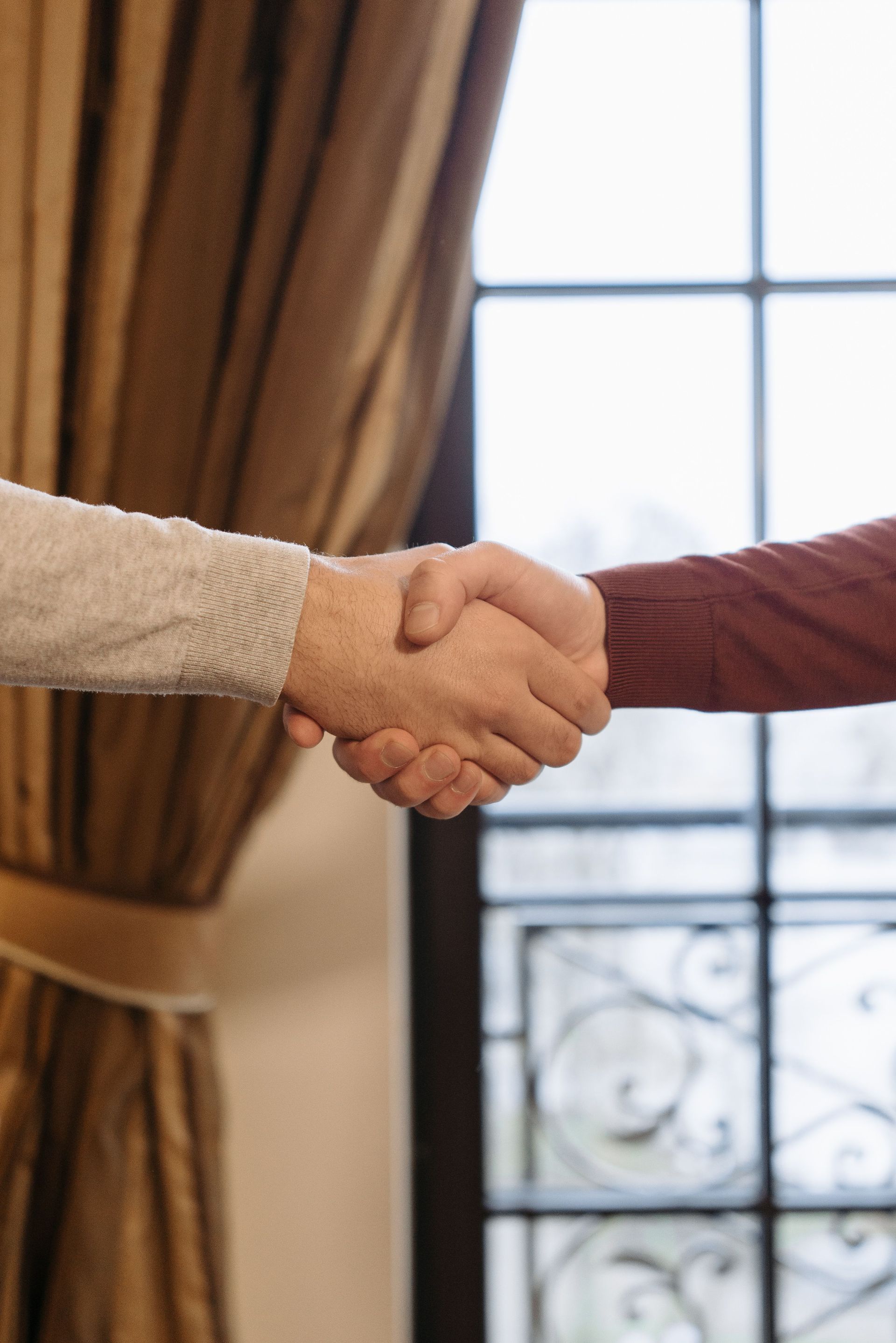 Two people shaking hands near a window with brown curtains