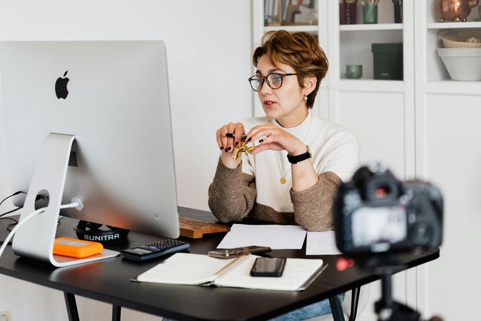 Woman with glasses speaks into a computer for a video, while a camera records from the foreground.