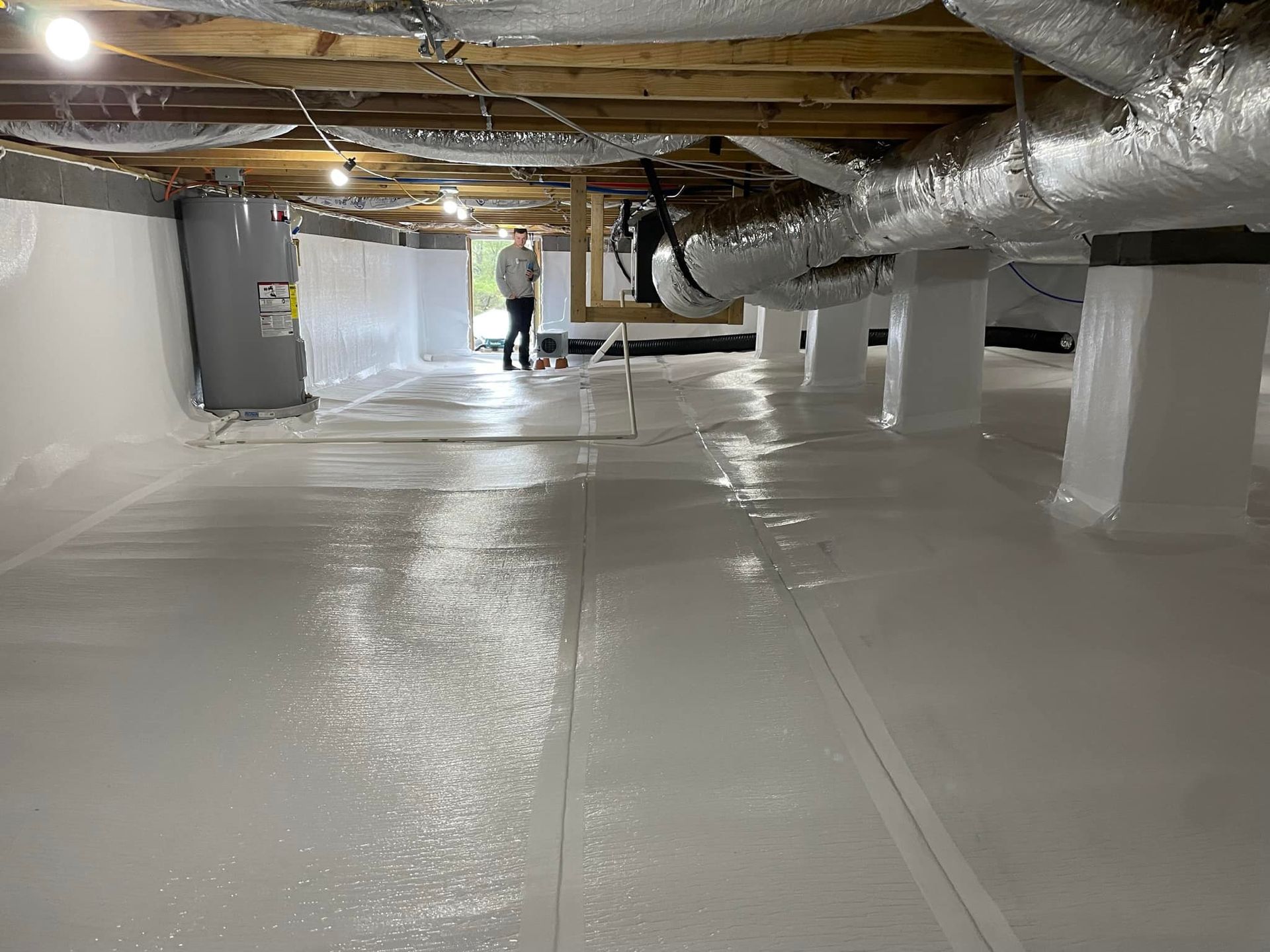 Person standing in a crawl space with white vapor barrier on walls and floor.