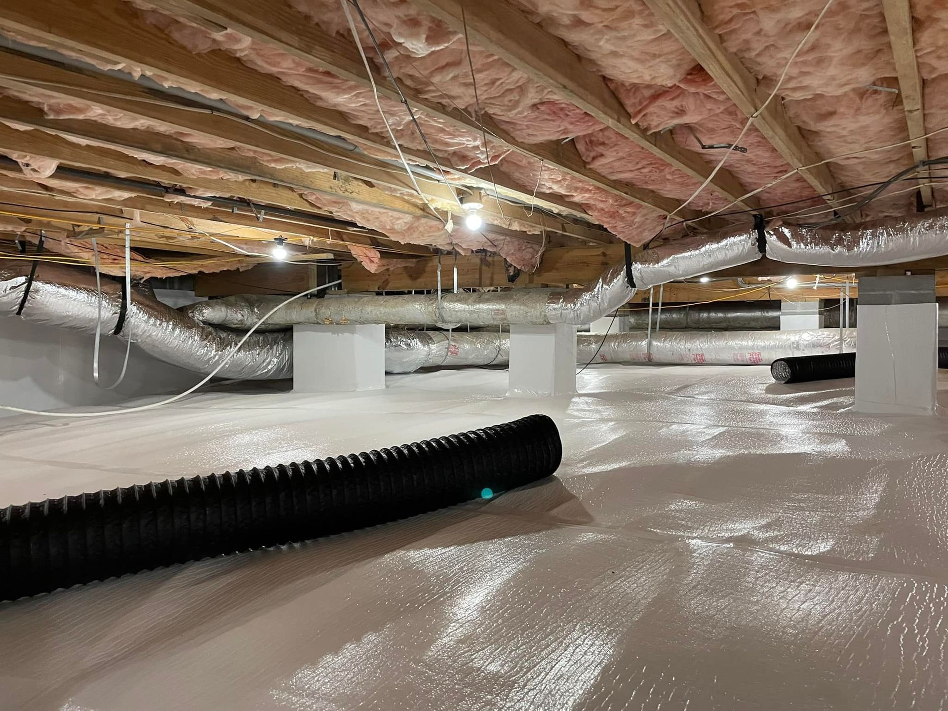 Crawl space with white vapor barrier on the ground, insulation overhead, and black flexible ducting.