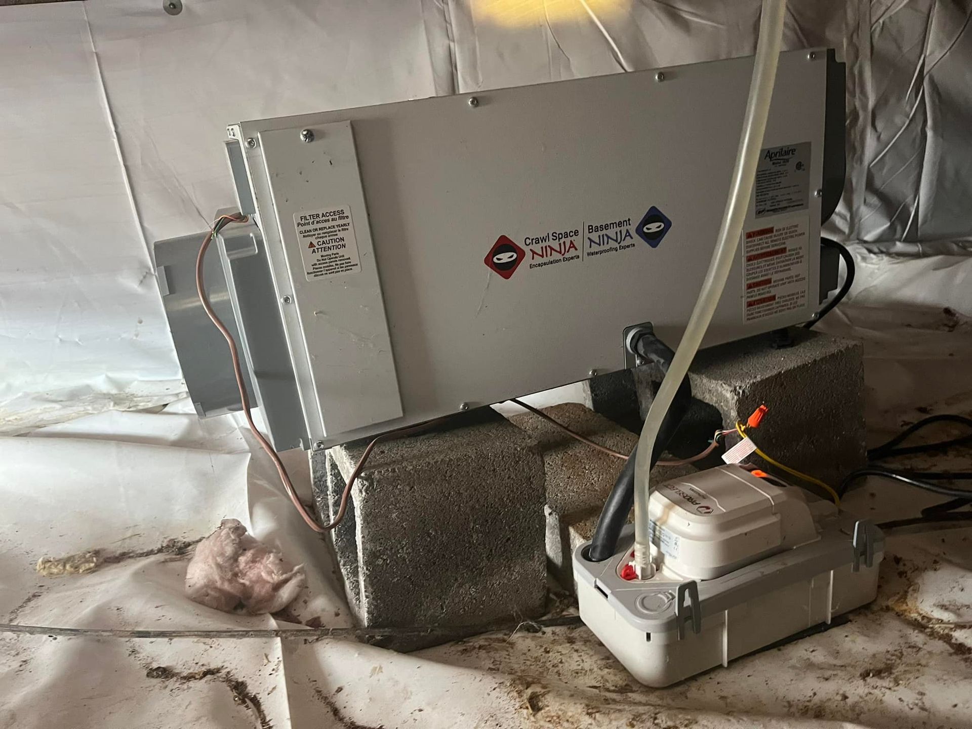Crawl space with a white dehumidifier, gray cinder blocks, and a pump; a white plastic vapor barrier covers the floor.