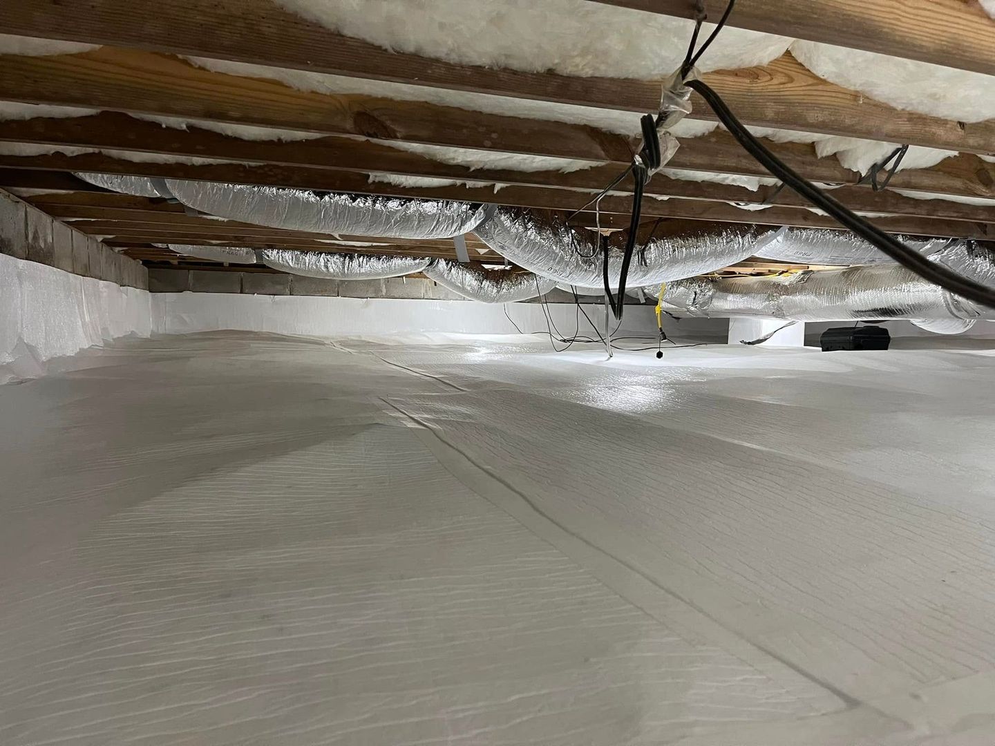 A crawl space with white insulation on the floor and walls, wood beams overhead.