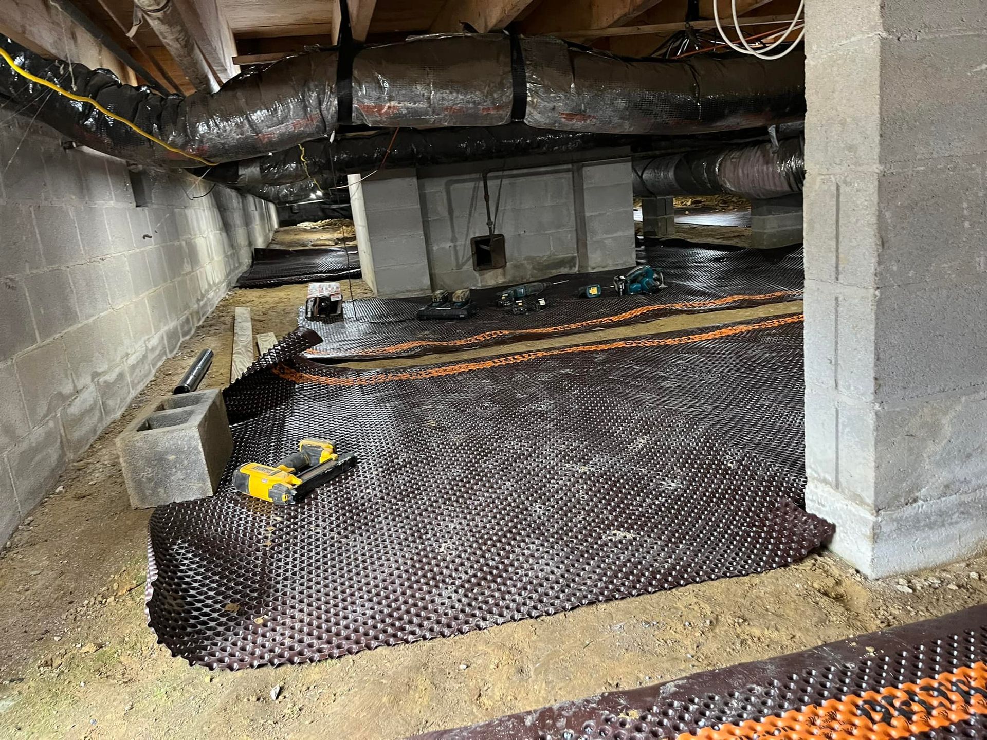 Under-house view with black drainage mat laid, concrete walls and supports, ducts, and a power tool.