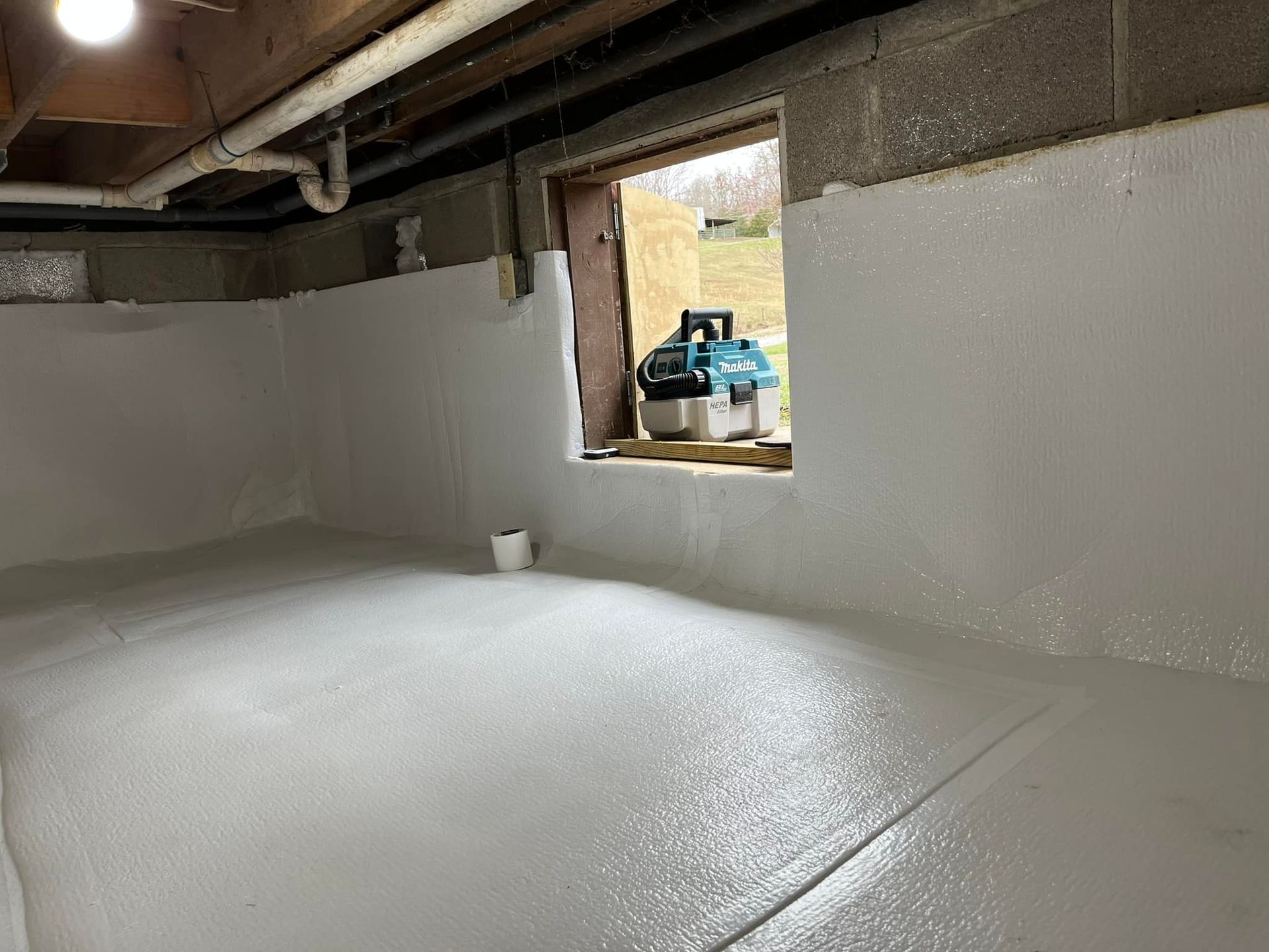 Basement with white spray foam insulation on walls and floor, window in the background with a tool.