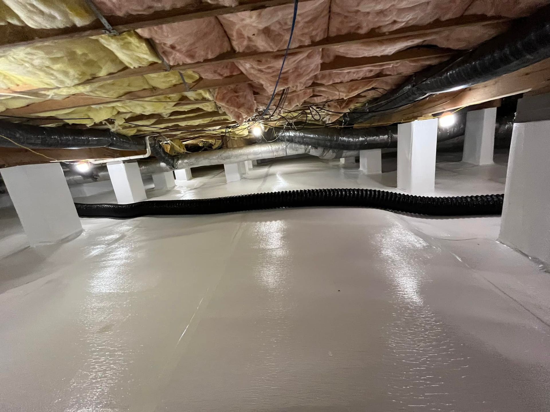 Crawl space with white floor, insulation, and support columns; black drainage system runs along the floor.