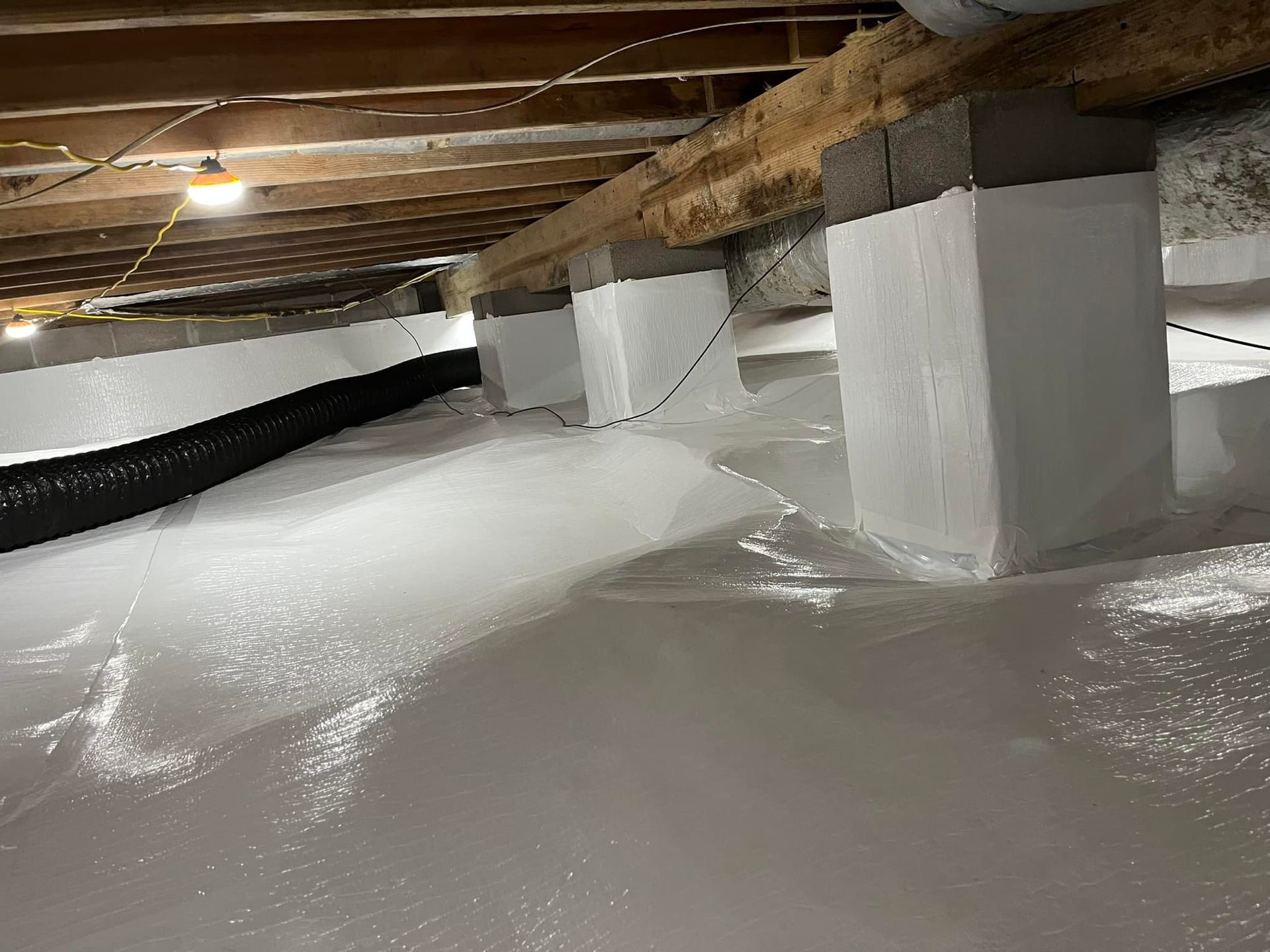 Crawlspace with white vapor barrier, support posts, and wooden beams. A black corrugated pipe is also present.