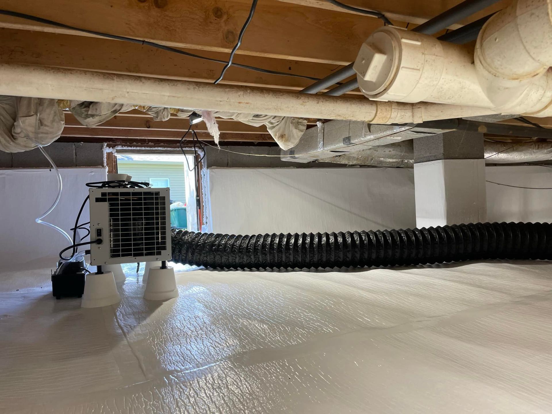 Crawl space with white plastic sheeting on the floor, ventilation system, and pipes under wooden beams.