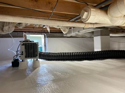 Crawl space with white plastic sheeting on the floor, ventilation system, and pipes under wooden beams.