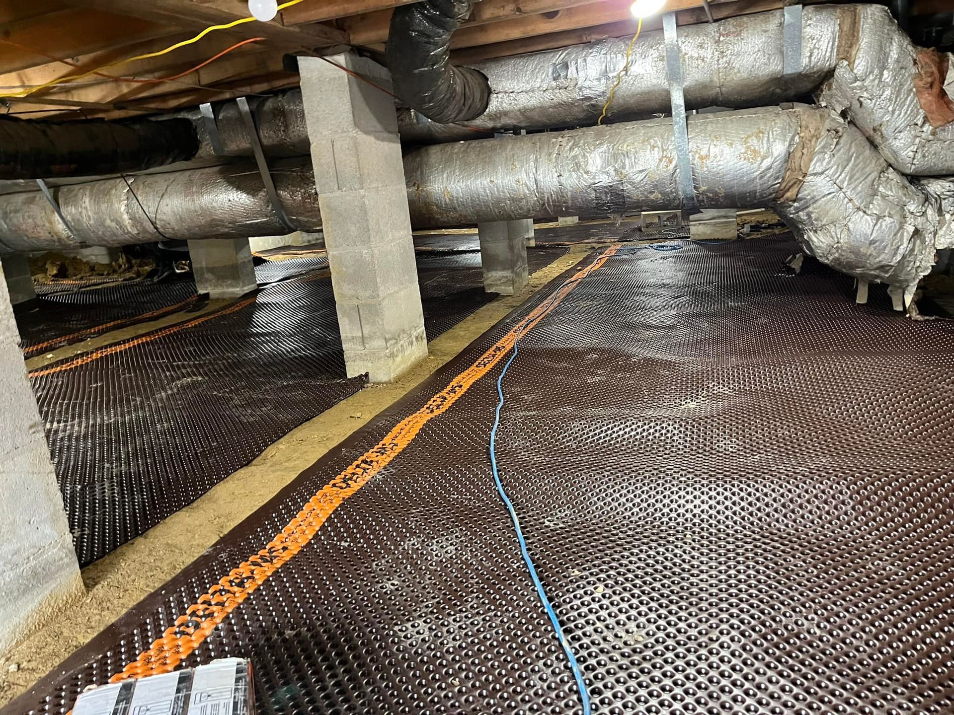 Black dimpled drainage mat in a crawl space, under pipes and supports.
