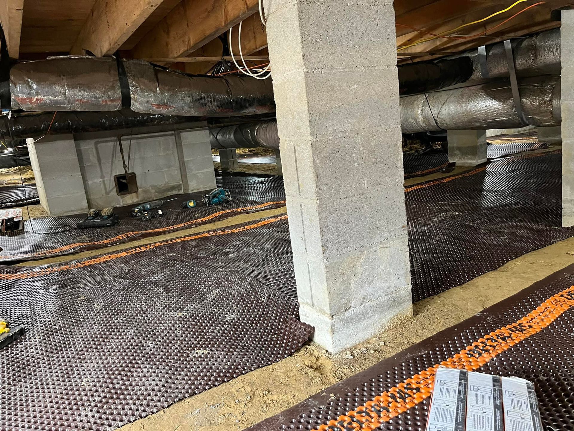 Underneath a building: concrete pillars, black and orange drainage mat, ductwork, and a damp environment.