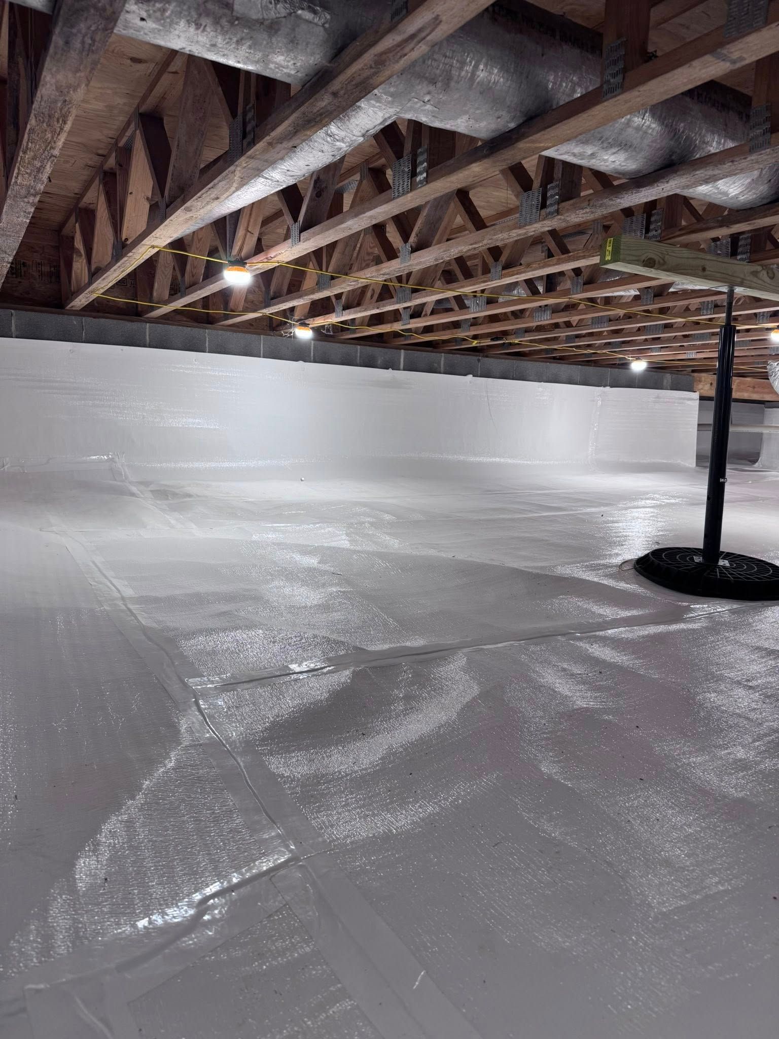 White vapor barrier covers a crawl space floor and walls; wood beams overhead.