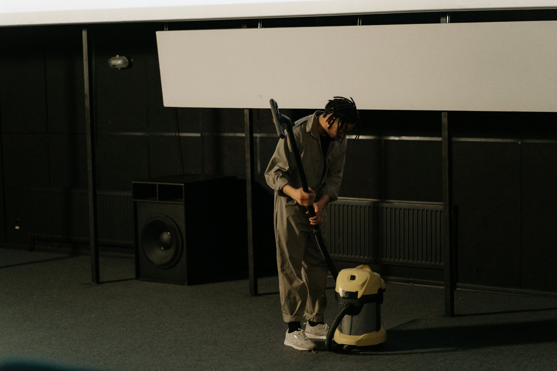 Person in grey jumpsuit vacuums a room. Black speaker, projector screen, and radiators in a dark space.