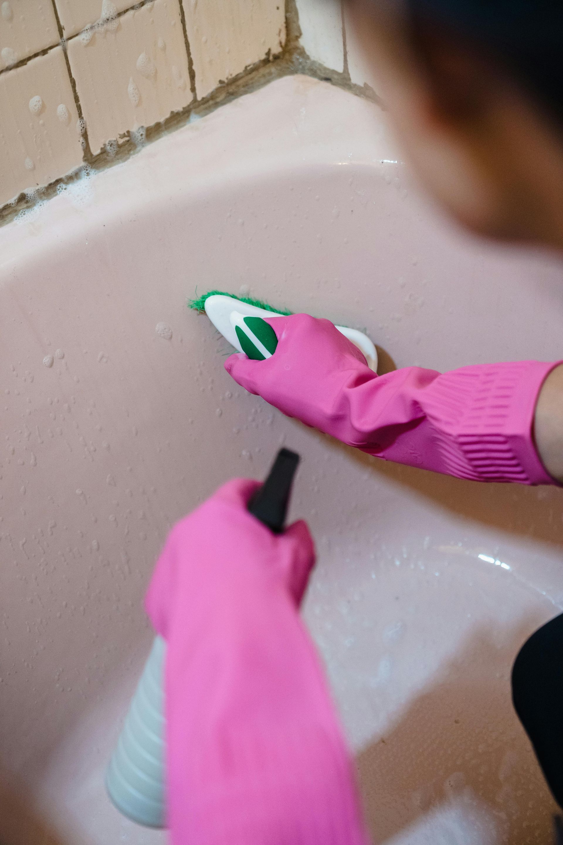 Person wearing pink gloves cleaning a pink bathtub with a scrub brush and spray bottle.
