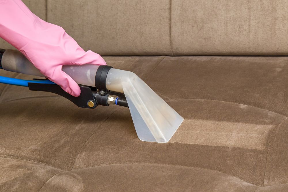 Person in pink gloves cleaning a brown couch with a steam cleaner.