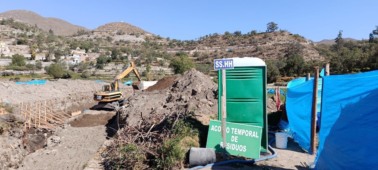 Un sitio de construcción con una excavadora, un baño portátil y montones de tierra cerca de una cadena montañosa.