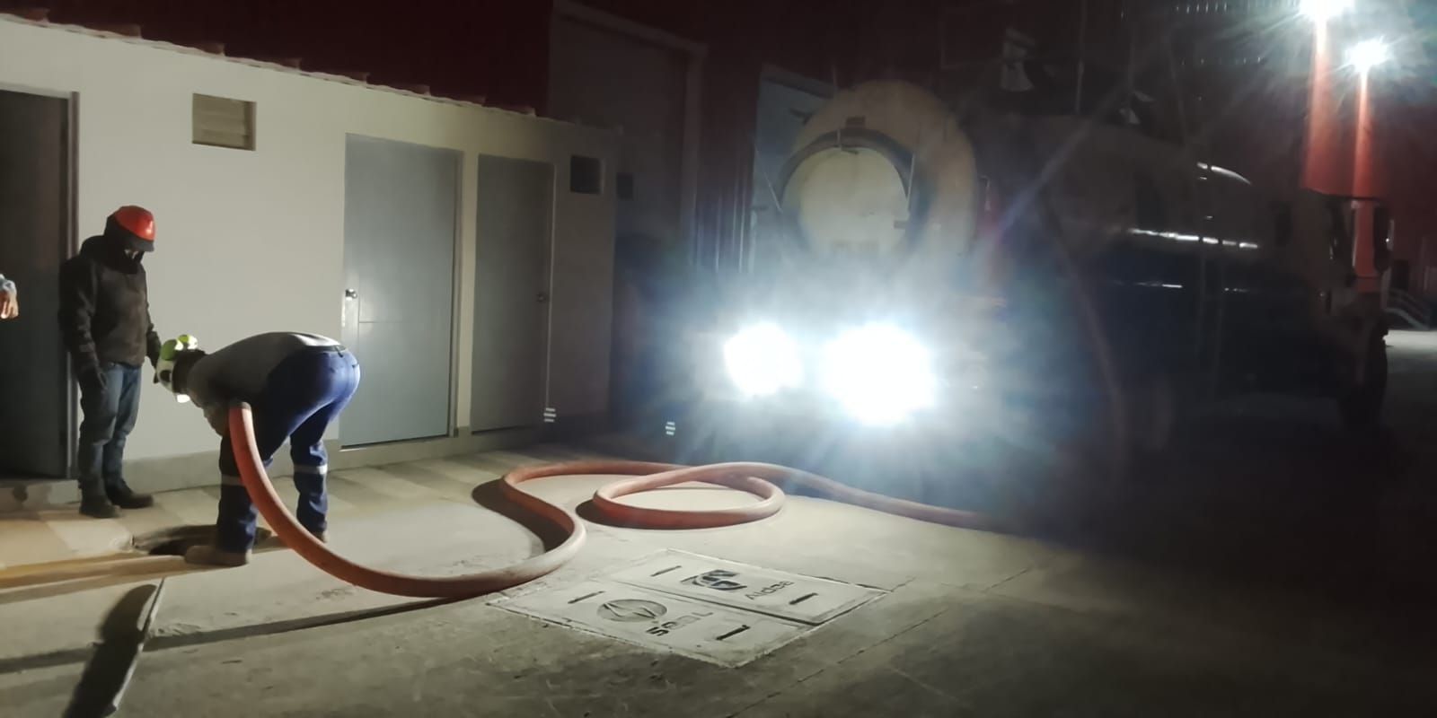 Bomberos conectando una manguera a un camión por la noche. Los faros brillantes iluminan la escena.