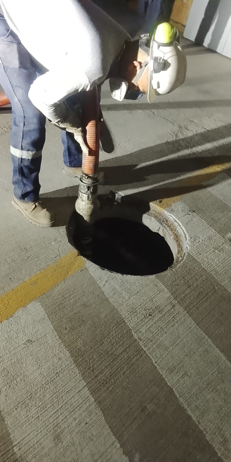 Trabajador vierte líquido oscuro en un agujero circular sobre una superficie de hormigón, vestido con ropa de trabajo y casco.