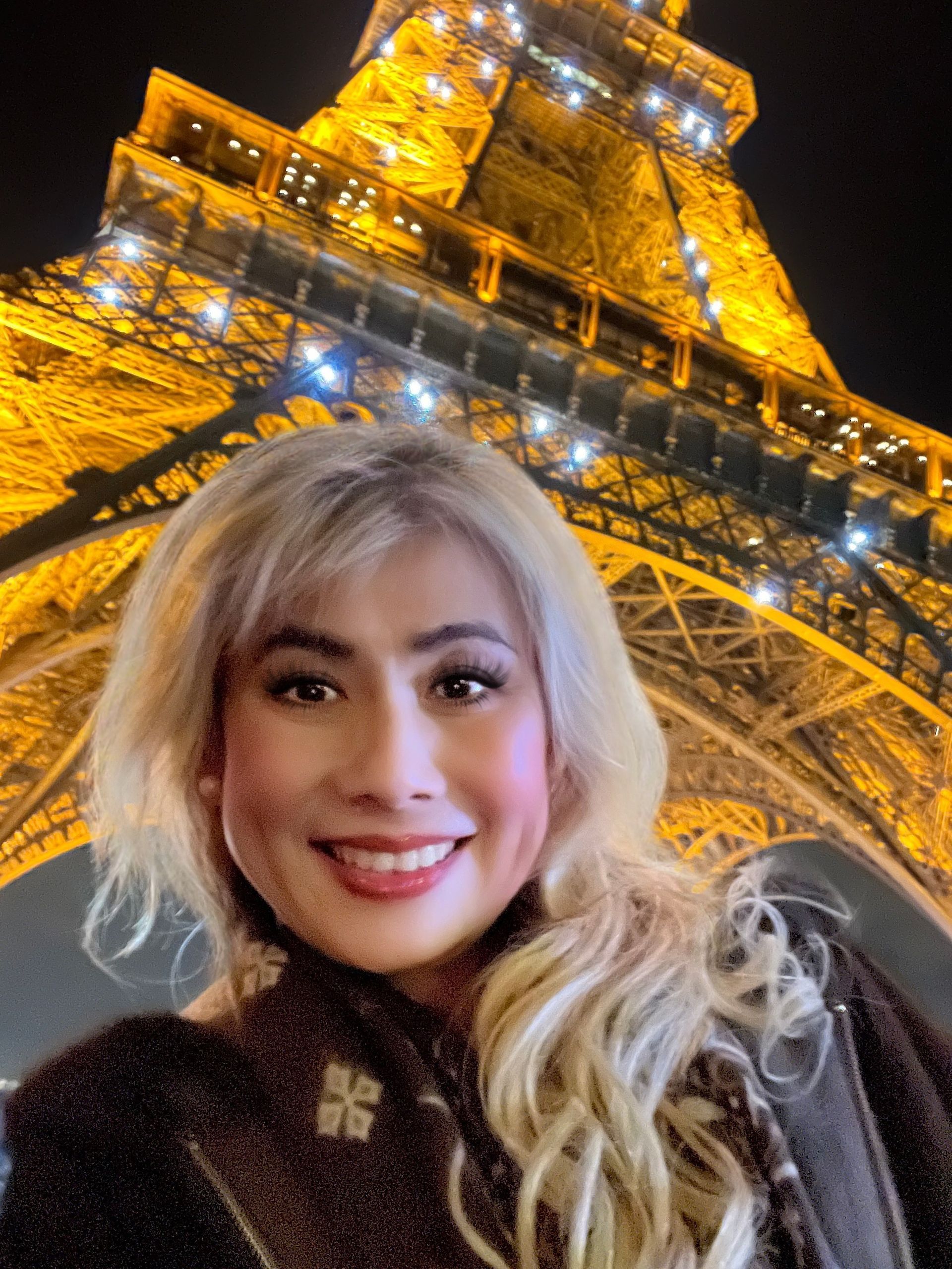 Woman smiling in front of the illuminated Eiffel Tower at night.