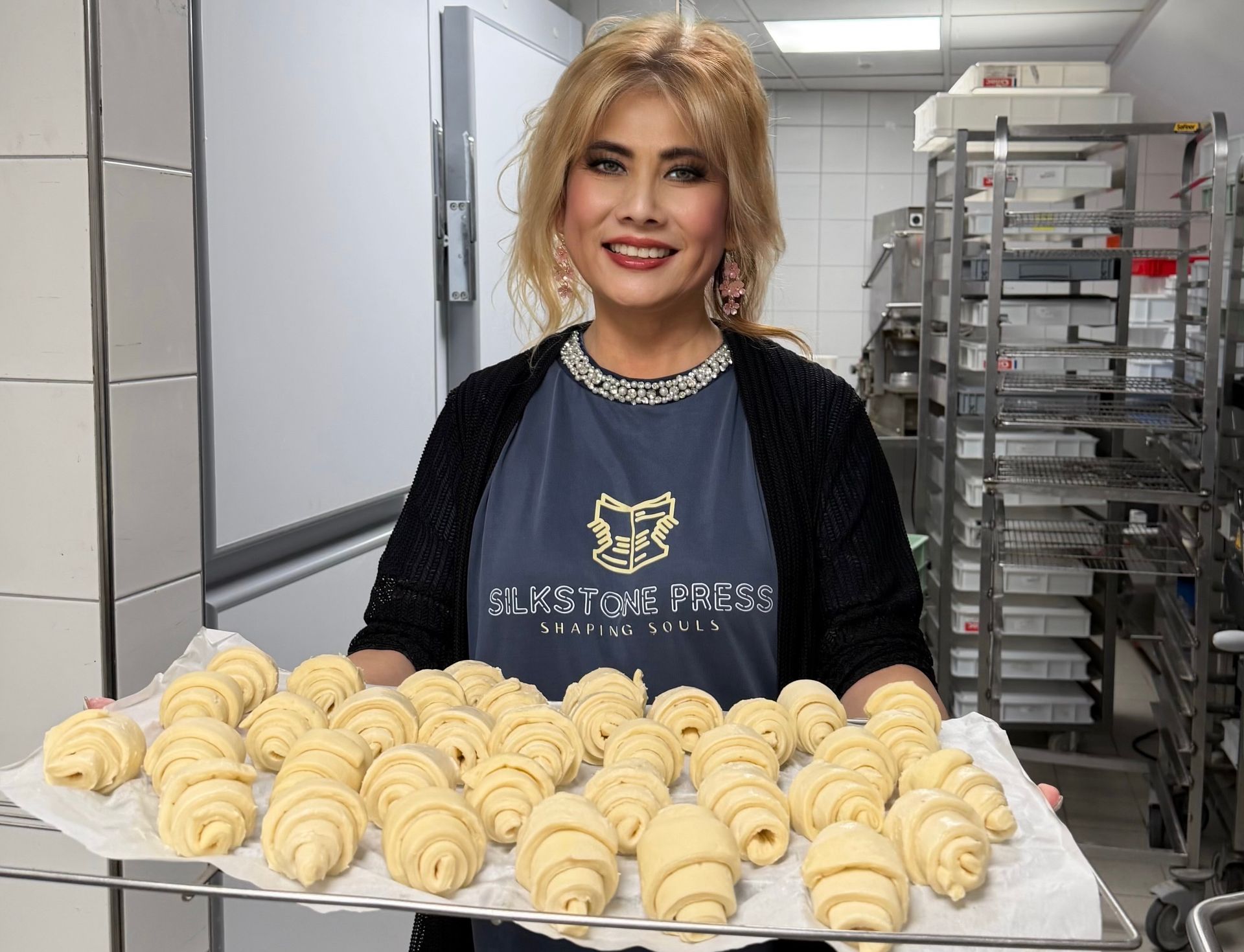 Woman holding a tray of raw croissants in a kitchen, smiling.