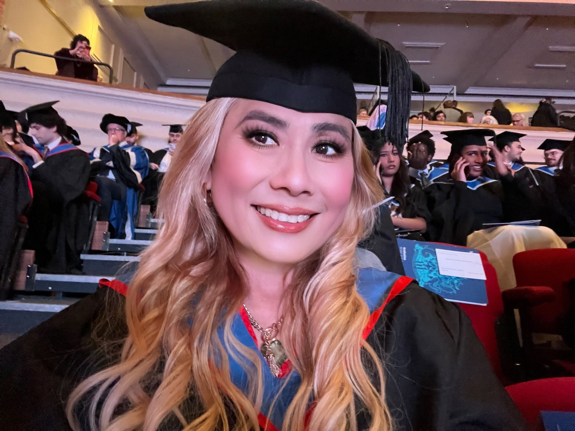 Woman in graduation attire smiles at camera in auditorium.