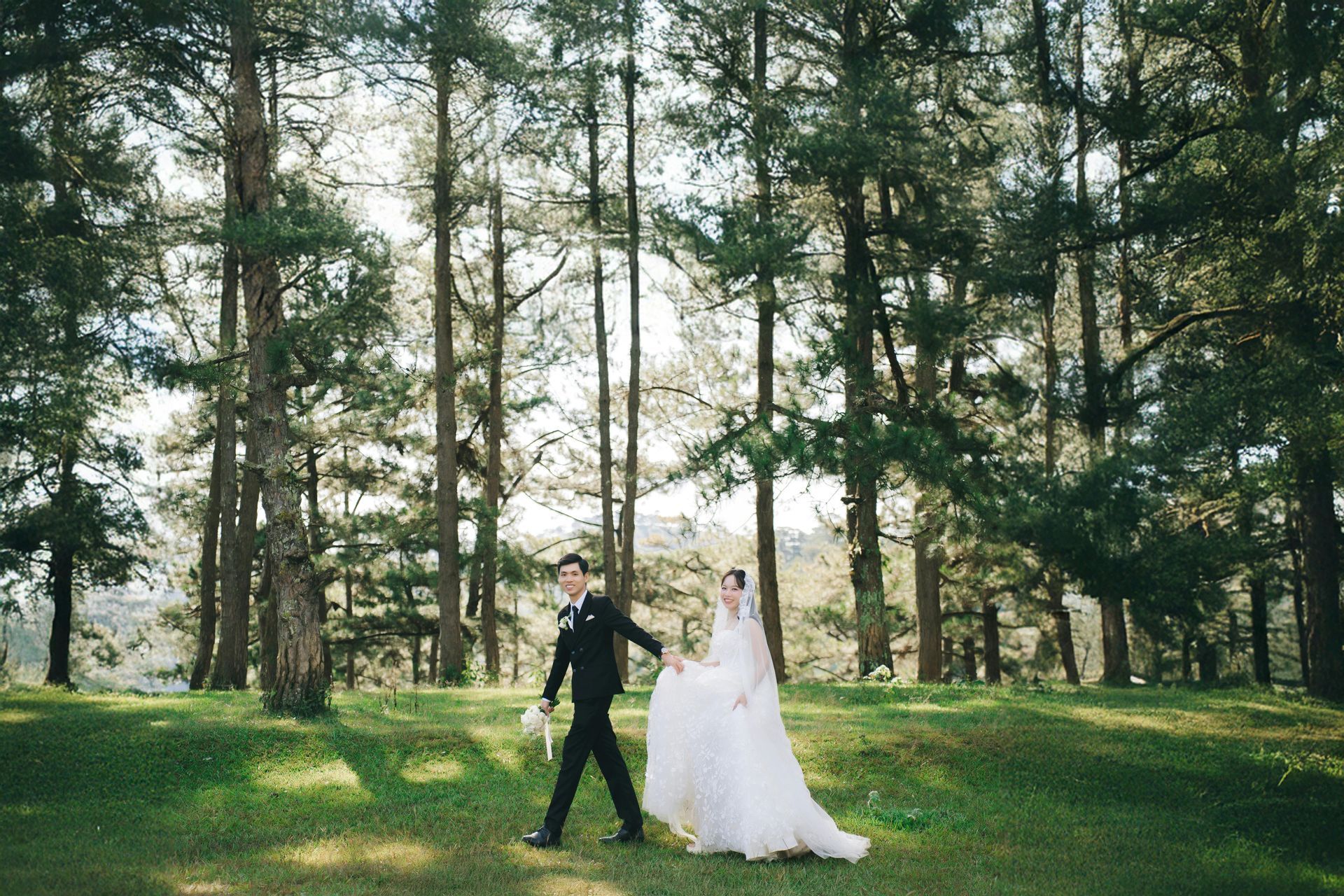 Couple in wedding attire walk through a sunny forest, groom holding flowers, bride's veil flowing.