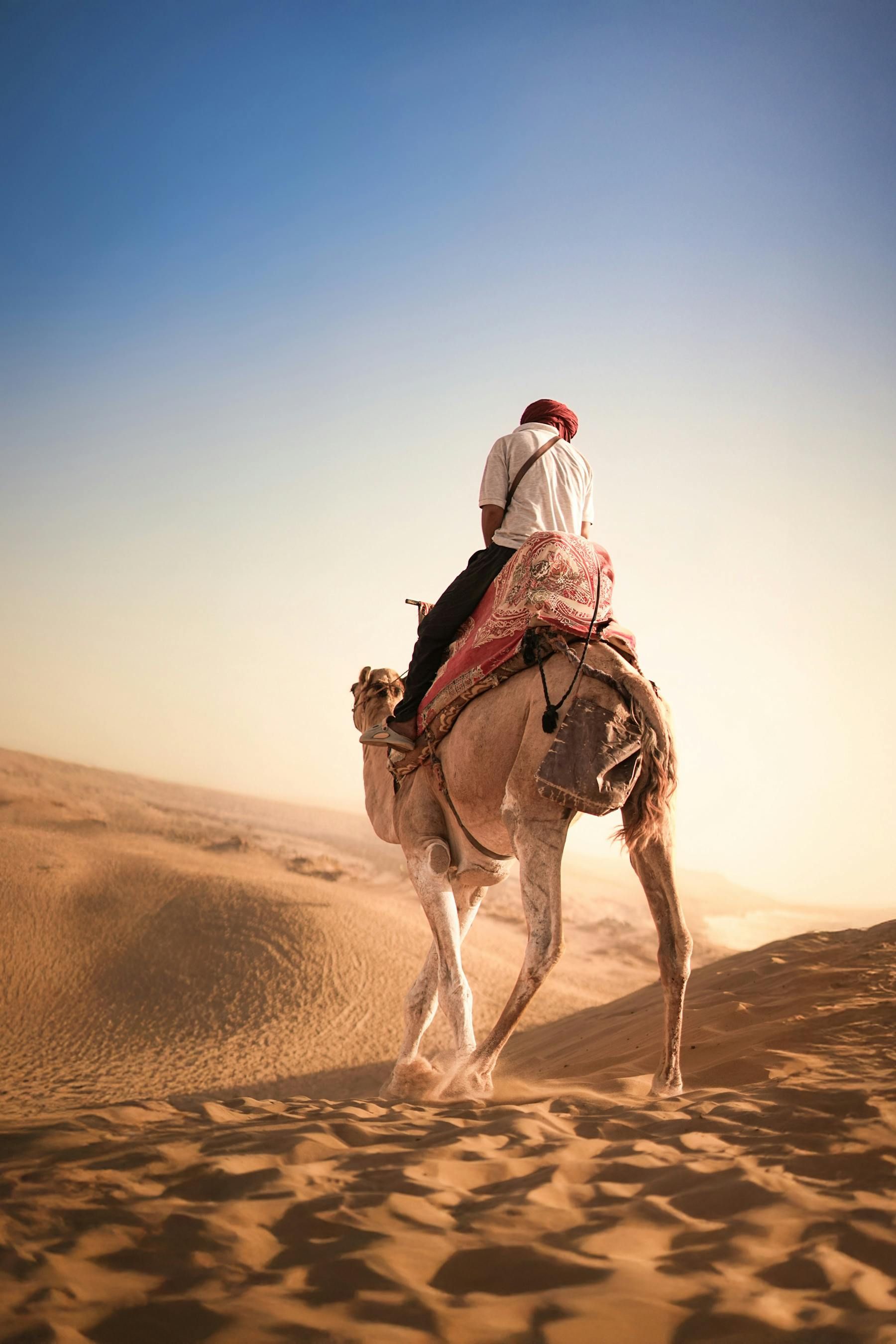 Man riding a camel through a desert, with sand dunes and a blue sky in the background.