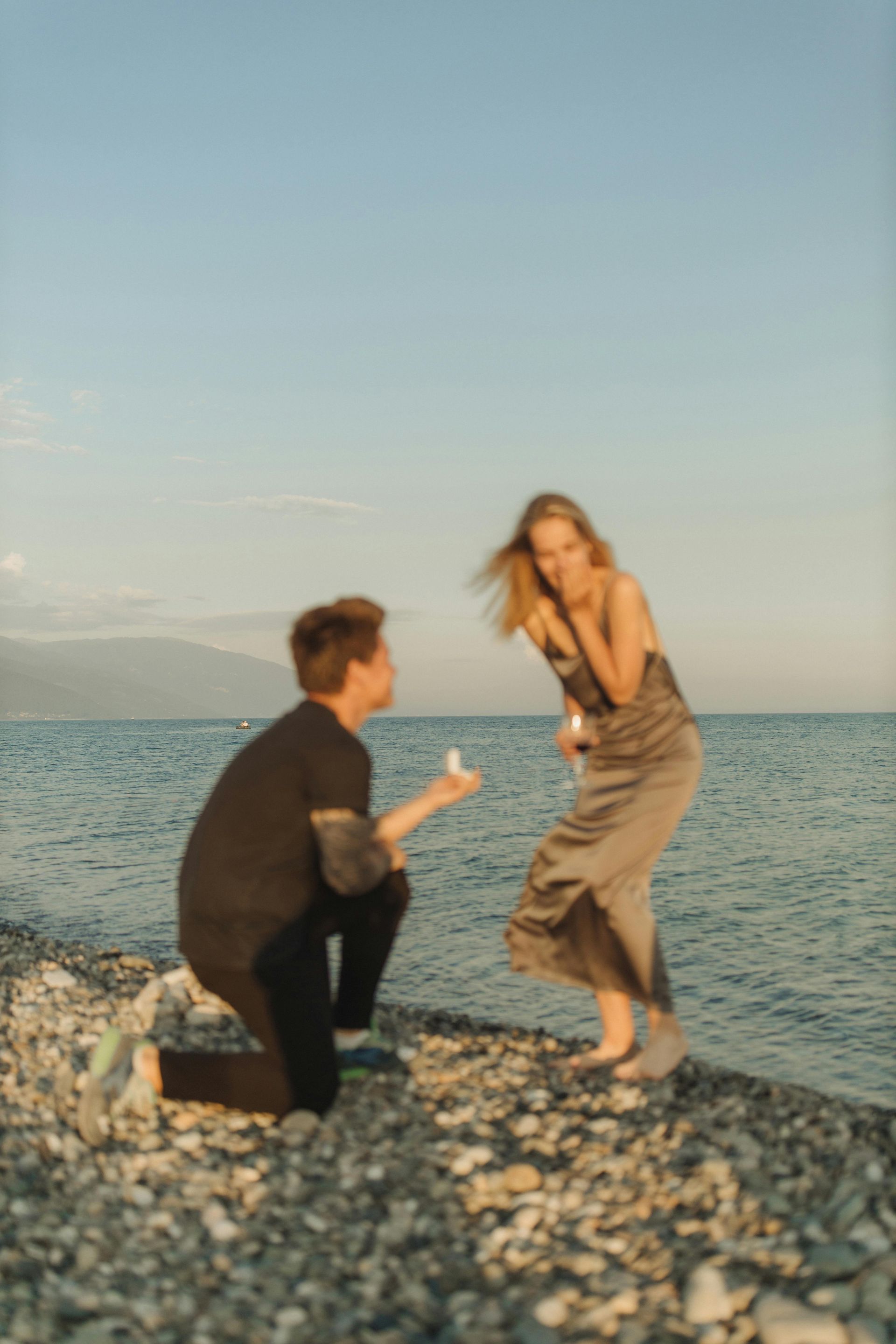 Person kneeling to propose on a pebble beach; woman reacts with surprise, ocean backdrop.