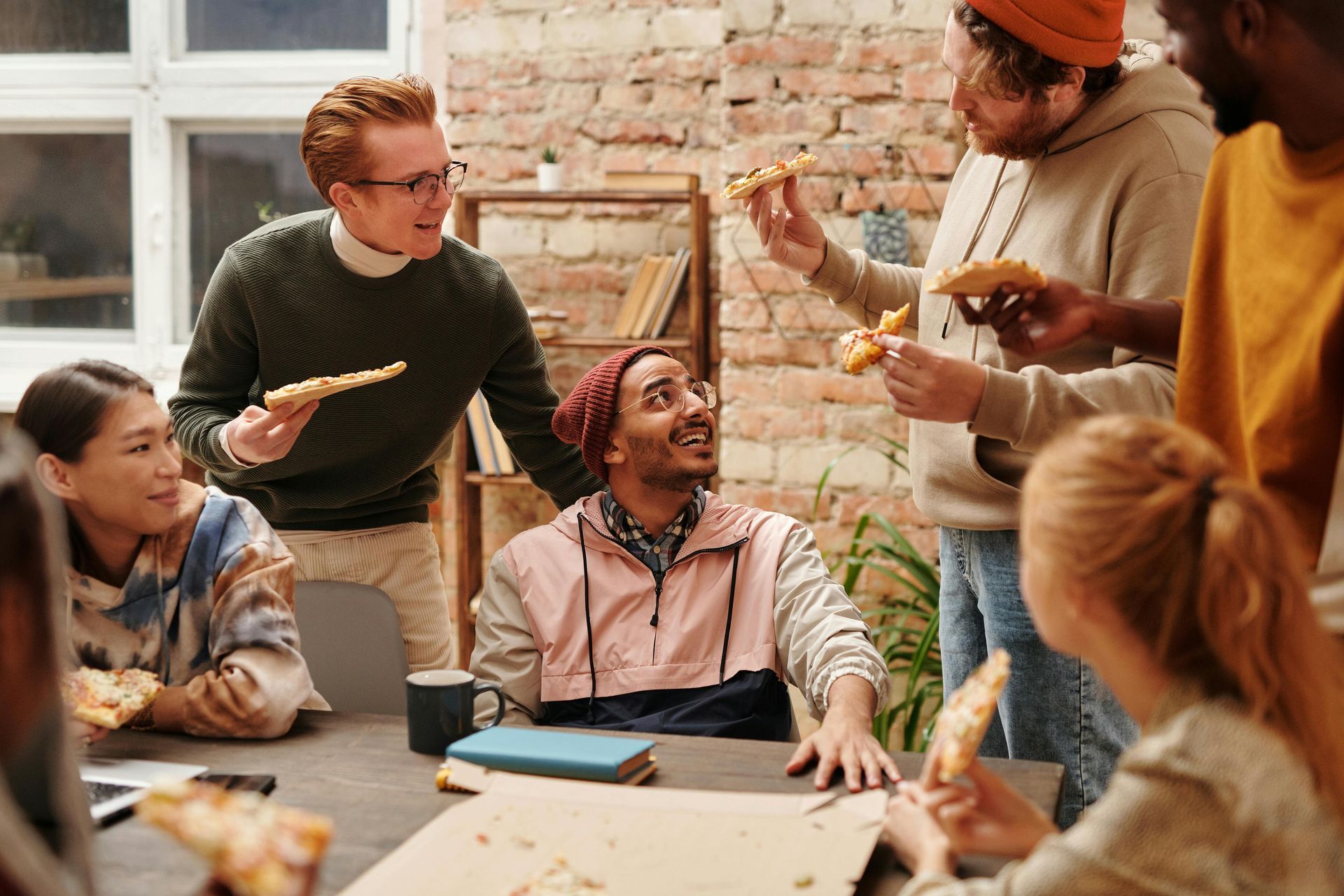 Group of diverse people enjoying pizza around a table. Smiling faces, casual clothing, exposed brick wall.