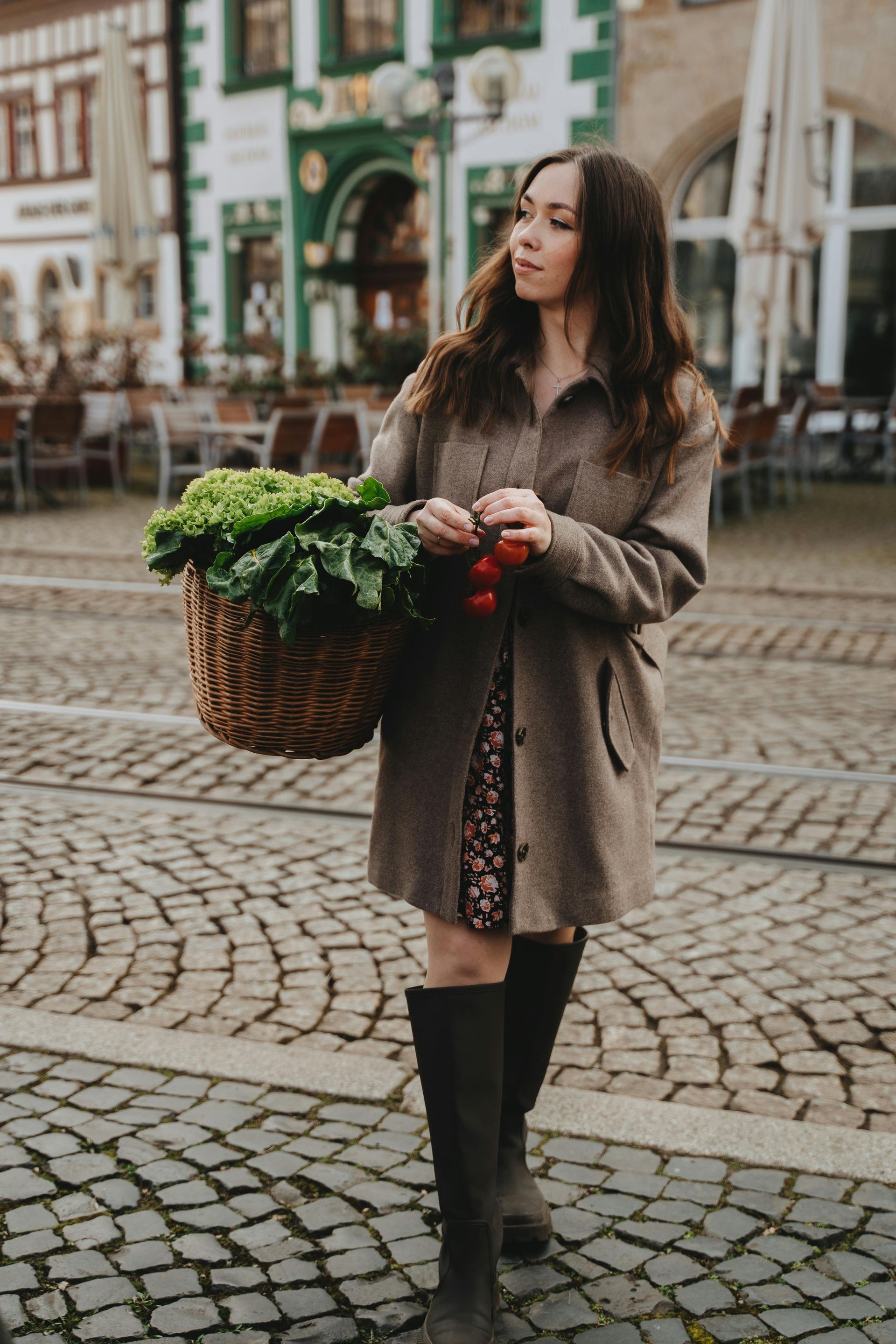 Woman holding a basket of vegetables, standing on a cobblestone street, wearing a brown coat and boots.
