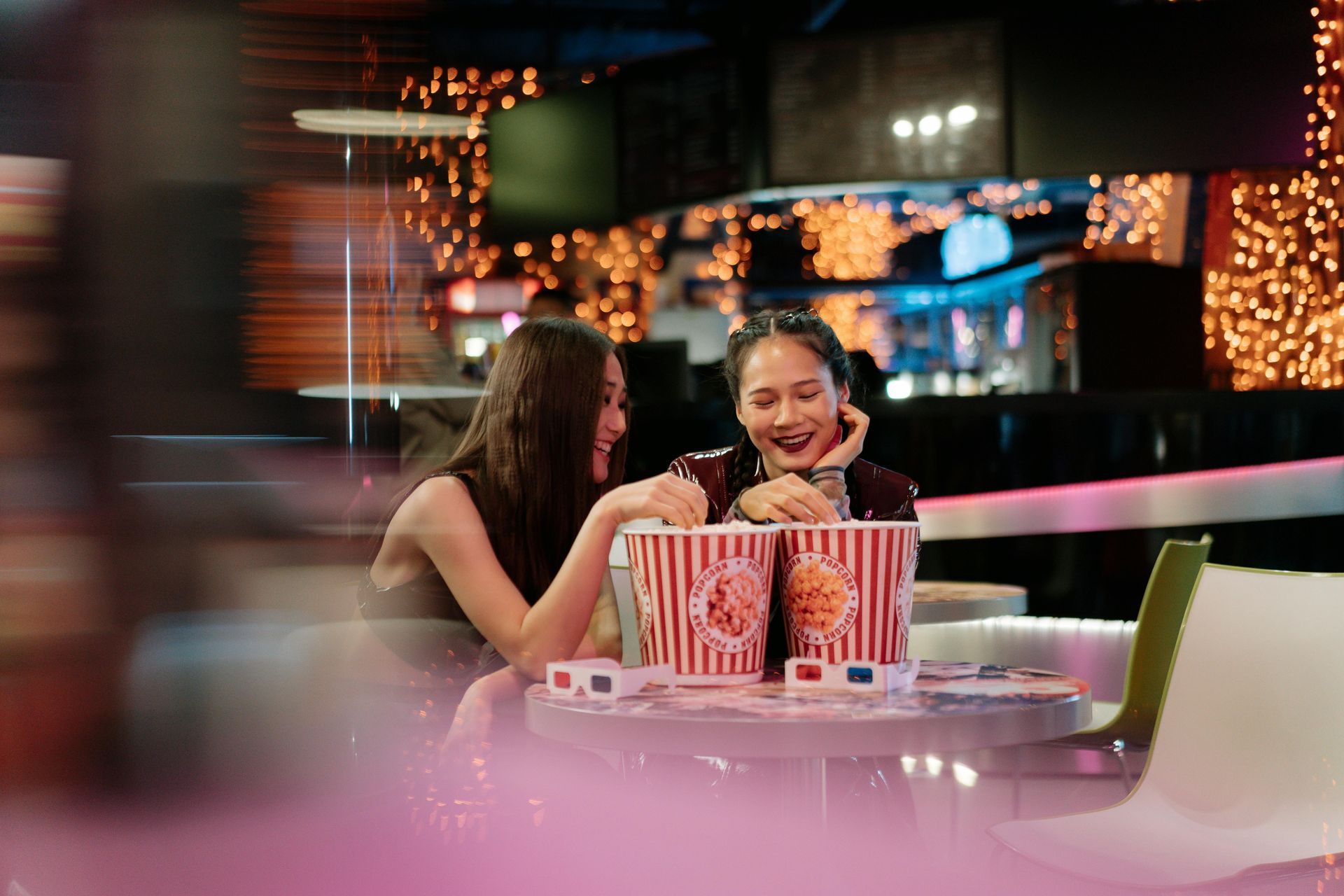 Two young women laugh, sharing popcorn at a movie theater table, 3D glasses nearby.