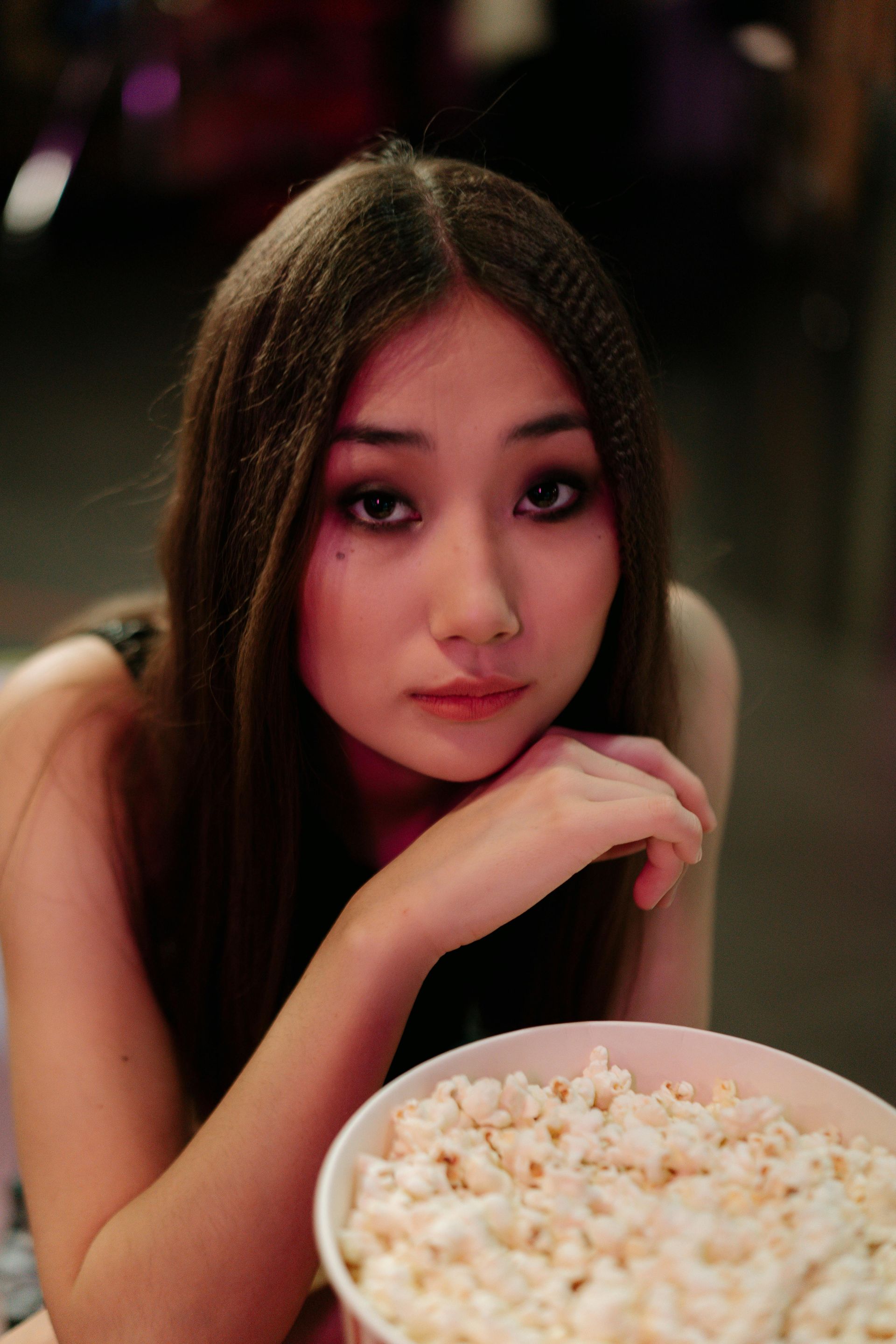 Woman with long crimped hair, holds a popcorn bucket, resting her chin on her hand and looking forward.
